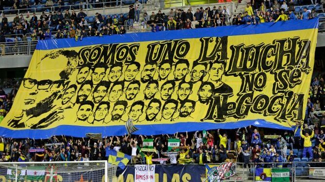 Aficionados despliegan una enorme pancarta con los rostros del entrenador y los jugadores para animar al equipo amarillo durante un partido.
