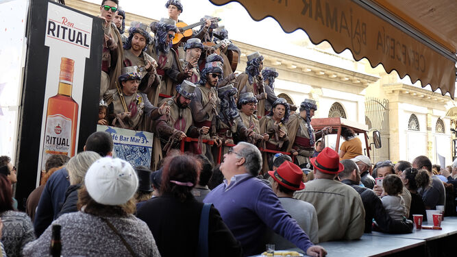Un grupo de personas consume en una barra de la calle, mientras presencia la actuación de un coro.
