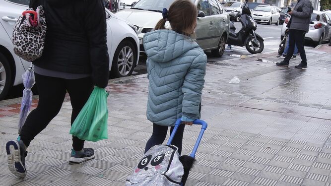 Una menor junto a su madre a la salida tira del carrito con su mochila a la salida del colegio.